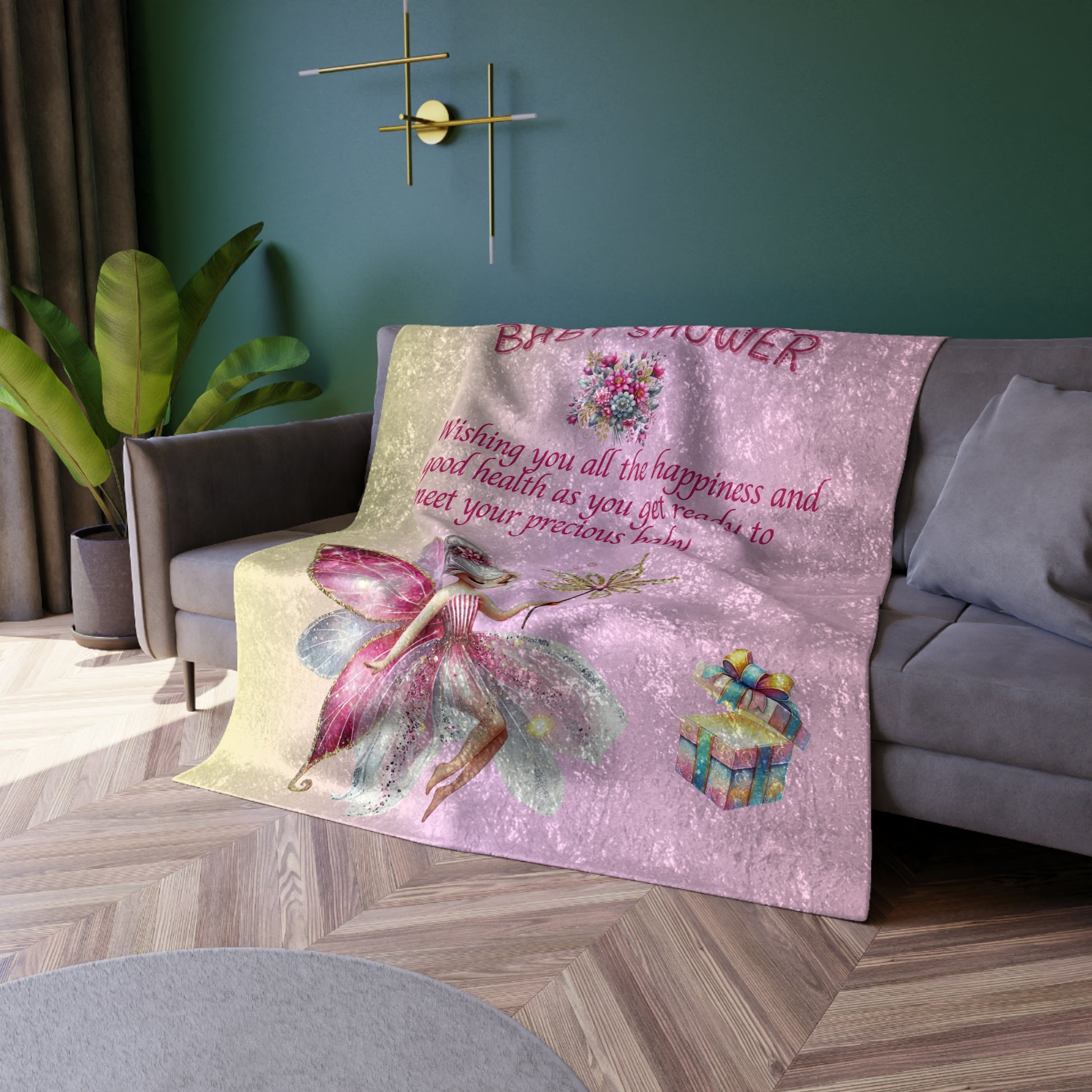 Pink baby shower blanket with fairy design on a gray sofa against a green wall.